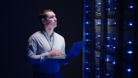 Young-man-holds-device-in-hand-and-looks-at-screen-inspects-equipment-or-hardware-rack.-Male-programmer-working-with-laptop-and-supporting-service-while-standing-in-data-center-spbas.